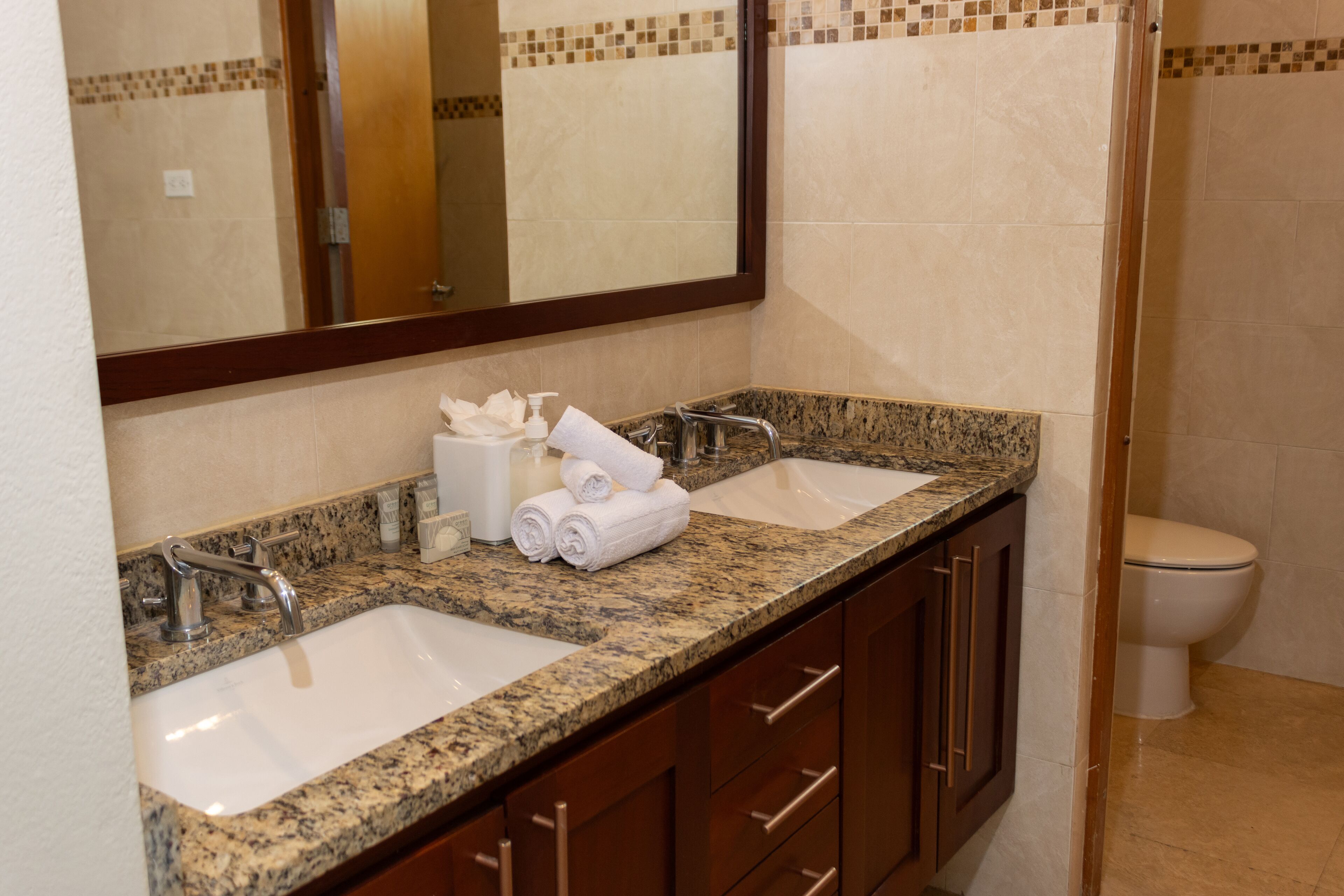 A bathroom with a double sink, a mirror, and a toilet.