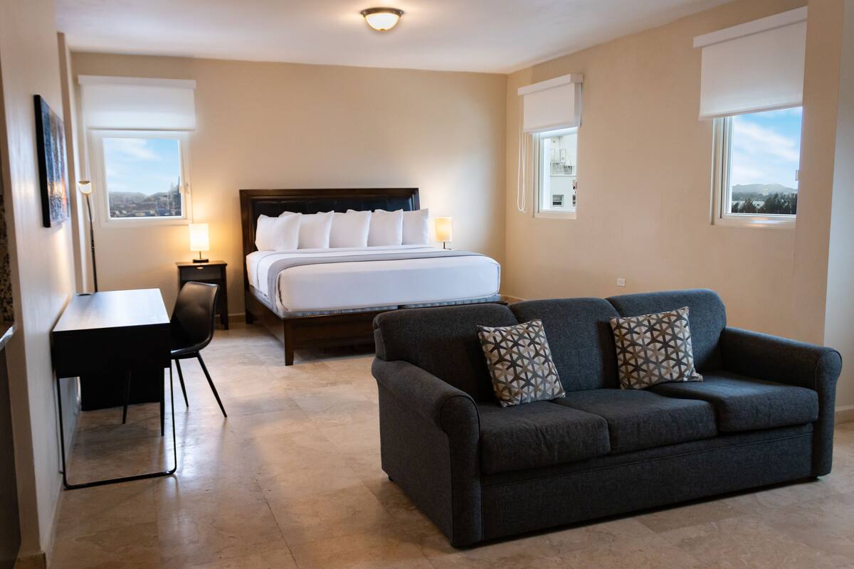 Habitación de hotel con cama, escritorio, sofá y dos ventanas con persianas.