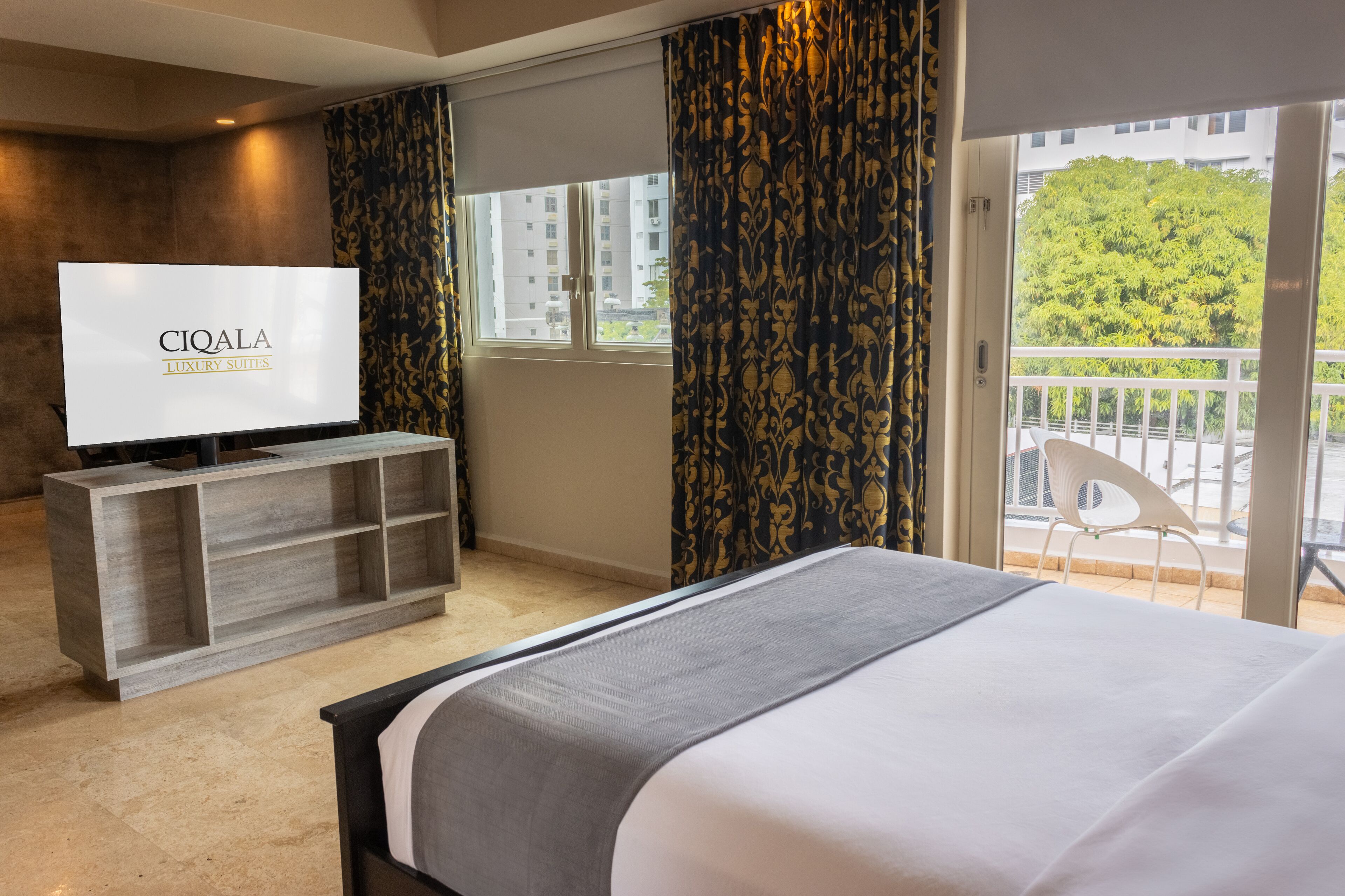 A hotel room with a bed, a TV on a wooden console, a balcony with a chair, and a view of buildings and trees.