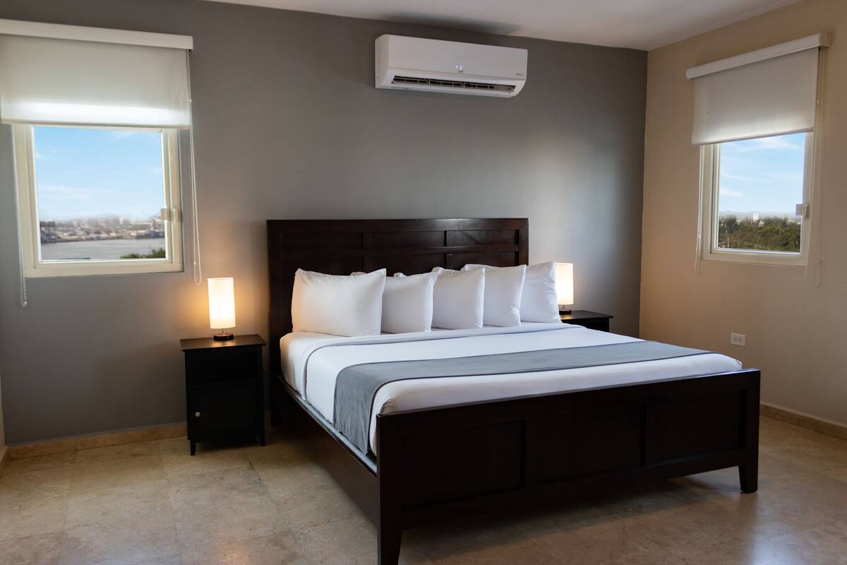 Un dormitorio con una cama de madera oscura, ropa de cama blanca, dos mesitas de noche con lámparas y vistas a la ciudad a través de ventanas.