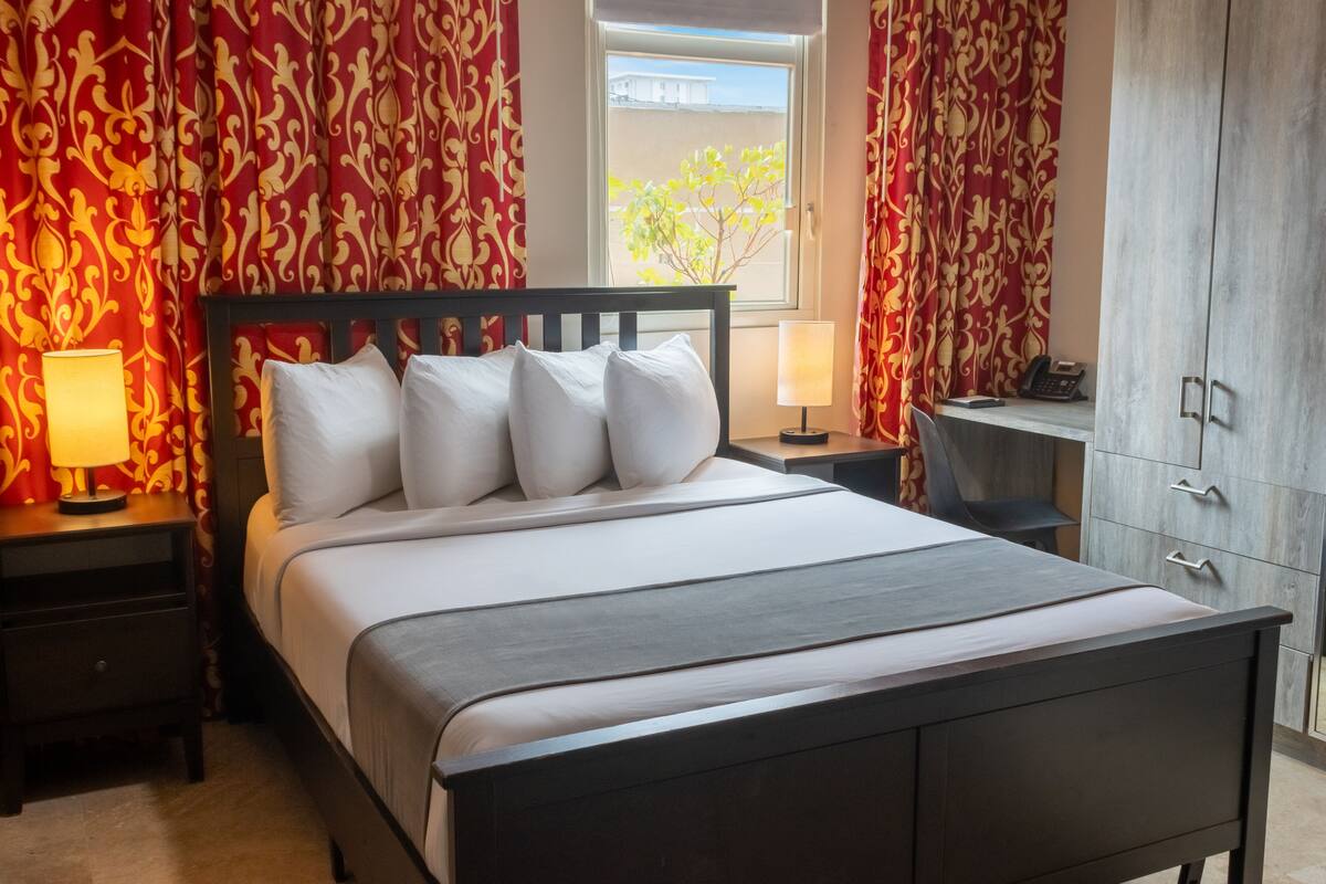 Una cama bien hecha con sábanas blancas y un cabecero oscuro, flanqueada por mesitas de noche con lámparas, en una habitación con cortinas rojas estampadas y vistas a través de una ventana.