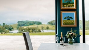 A table with a bottle of champagne, a glass, and a vase of flowers, set against a scenic view of a lake and hills. - The Coniston Hotel and Country Estate (Skipton)