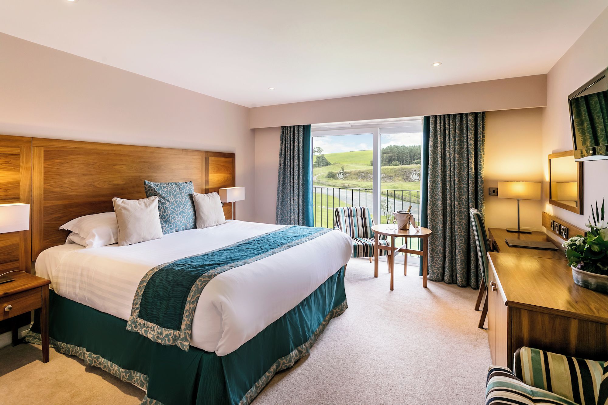 A hotel room with a large bed, a desk, a chair, a TV, and a balcony with a view.