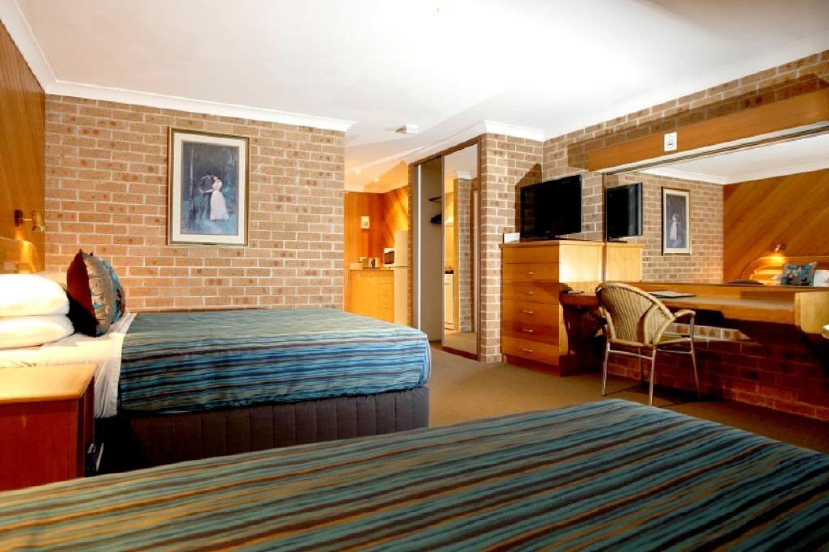 A hotel room with a brick wall, a bed with a striped bedspread, a desk with a chair, a television, and a framed picture on the wall.