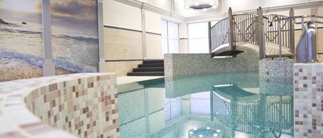 An indoor swimming pool with a mosaic tile design, a staircase with a glass railing, and a large window with a beach view.