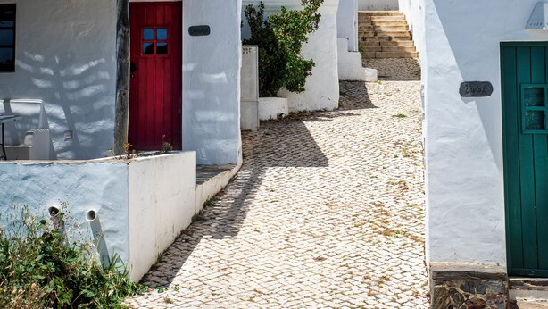 Une ruelle étroite pavée, bordée de bâtiments blancs aux portes rouges et vertes, sous un ciel bleu dégagé.