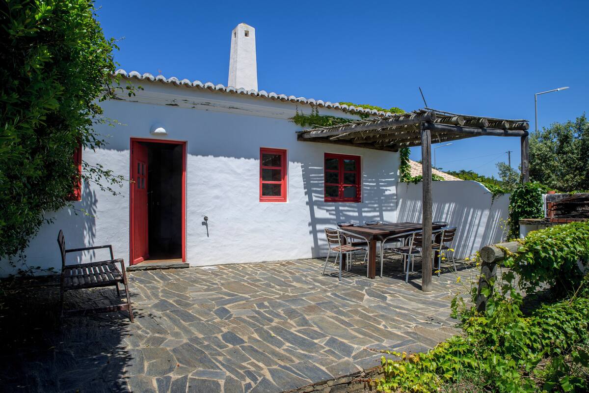 A white building with red doors and windows, a stone patio, and a wooden table and chairs.