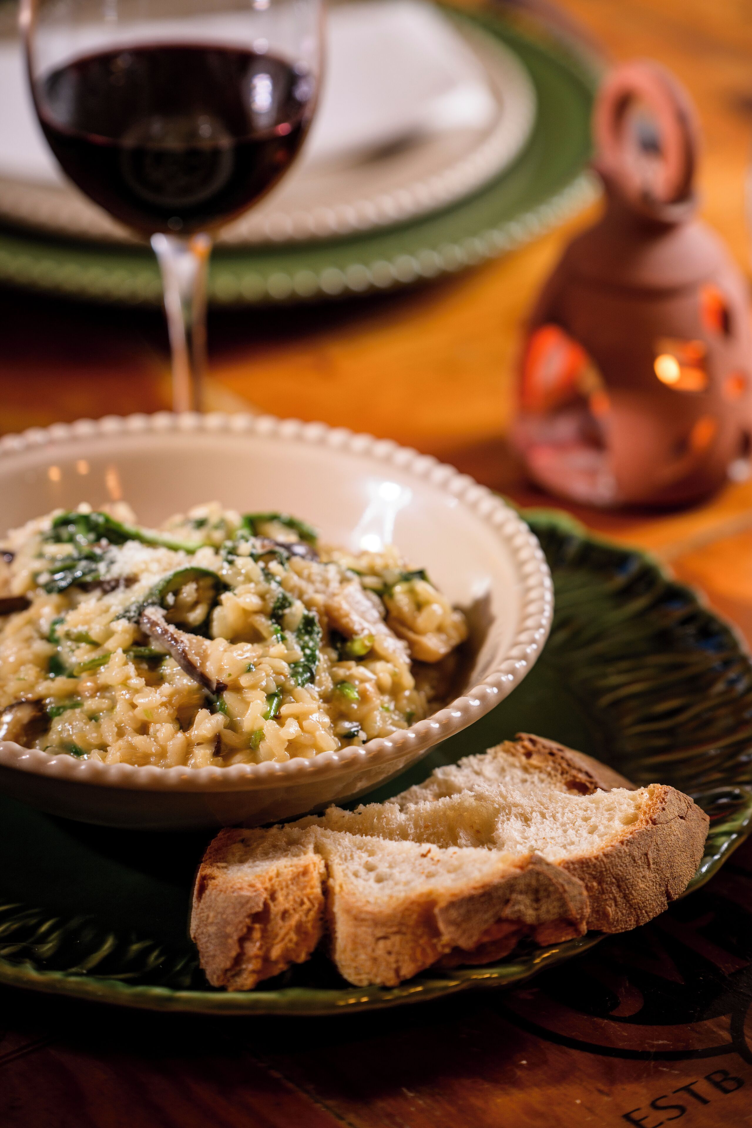 Un tazón de risotto con champiñones y espinacas, una copa de vino tinto y una rebanada de pan sobre un plato verde.