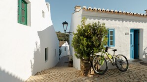 Une ruelle étroite pavée, avec un bâtiment blanc orné de portes et de fenêtres bleues, un vélo garé devant et un petit arbre.