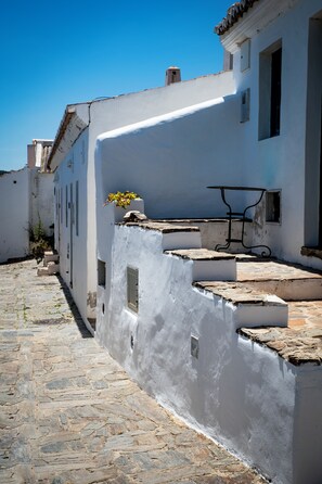A narrow cobblestone street with white buildings, a bench, and a small flower pot.