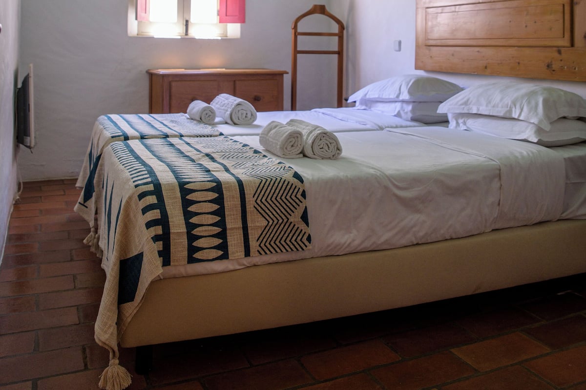 A bedroom with a bed, a window with shutters, a wooden headboard, and a brick floor.