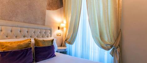 A hotel room with a tufted headboard, a bedside table with a lamp, and sheer curtains.