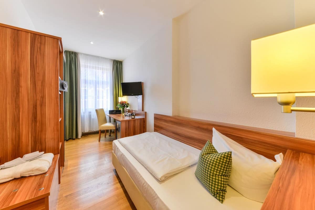 Habitación de hotel con un armario de madera, un escritorio con silla, una cama con almohada verde a cuadros y un televisor fijado a la pared.