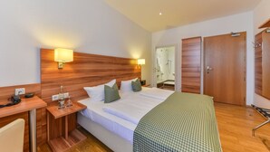 Habitación de hotel con cabecera de madera, cama con sábanas blancas, mesita de noche con lámpara, escritorio con teléfono y baño visible a través de una puerta abierta.