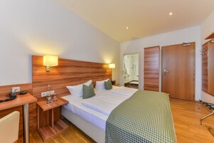 Habitación de hotel con cabecera de madera, cama con sábanas blancas, mesita de noche con lámpara, escritorio con teléfono y baño visible a través de una puerta abierta.