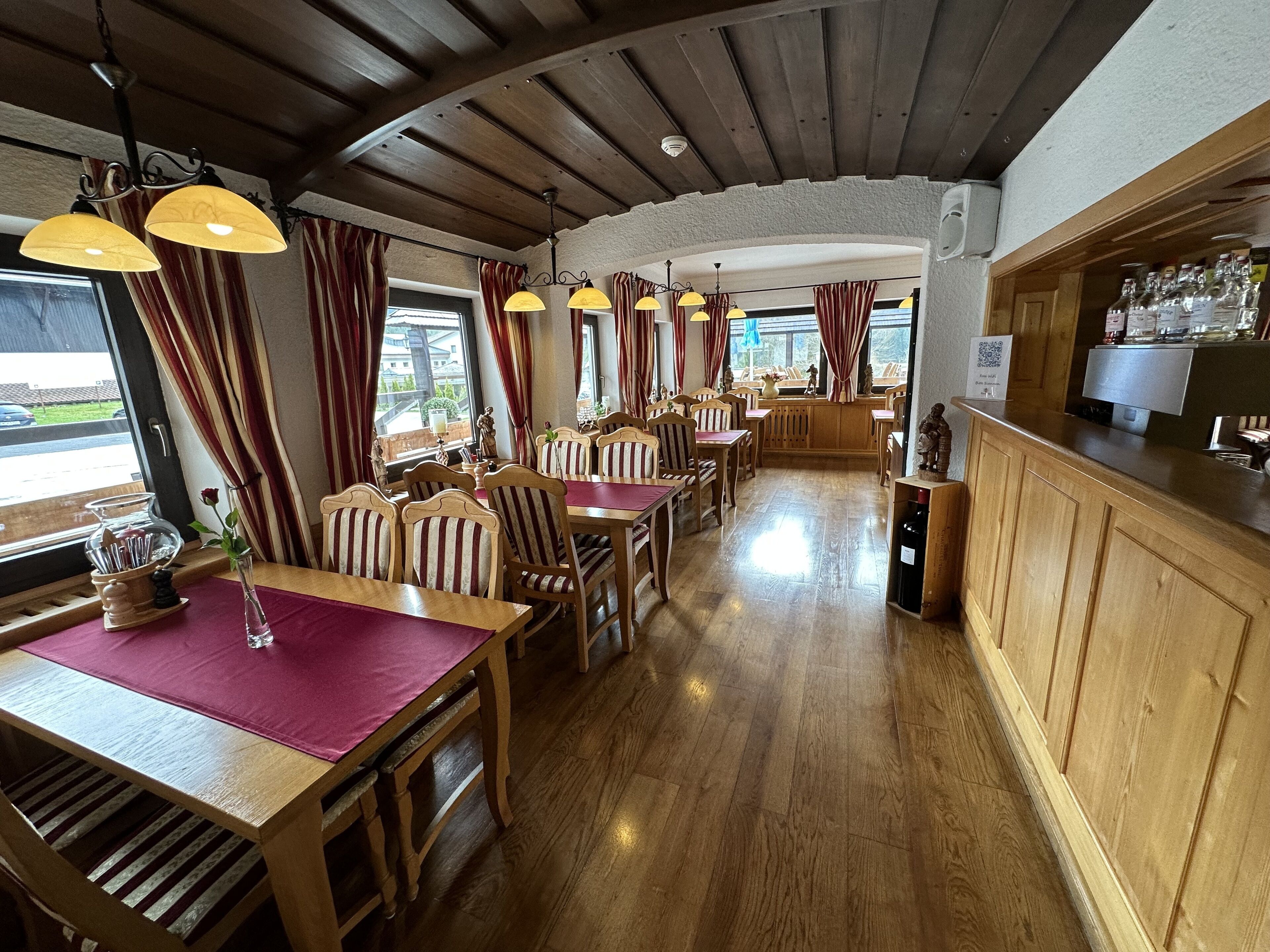 Un restaurante con mesas y sillas de madera, una zona de bar y amplios ventanales con cortinas.