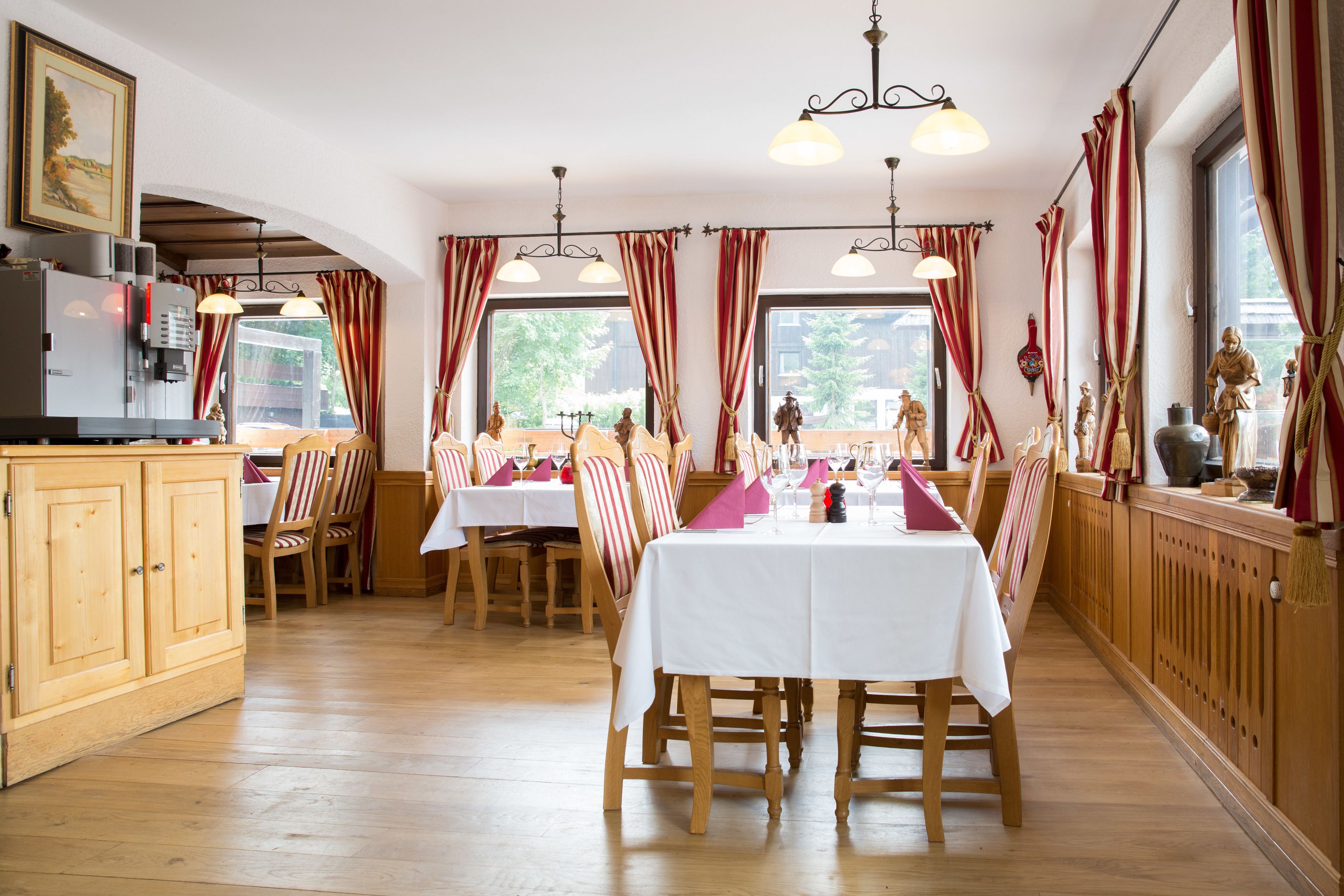 Un comedor con mesas y sillas de madera, una zona de cocina con refrigerador y ventanas grandes con cortinas rojas.