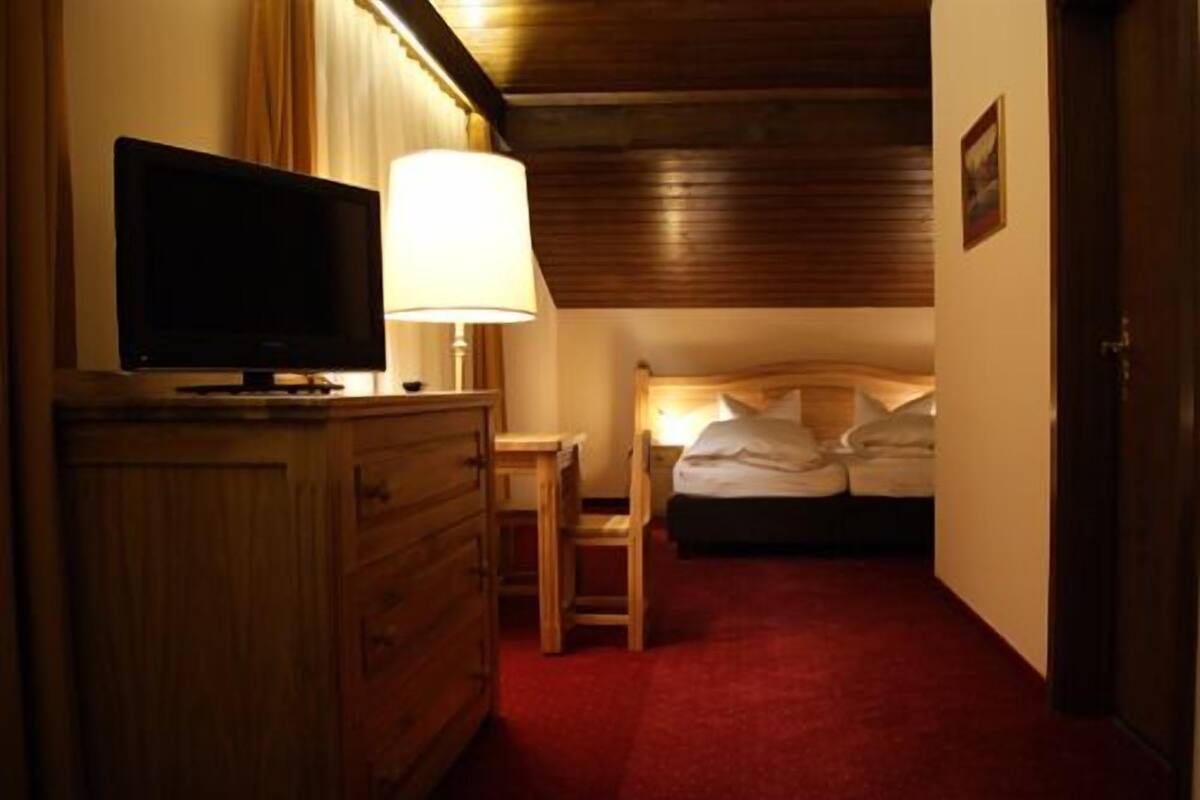 A hotel room with a wooden dresser, a television, a bedside table with a lamp, and a bed with pillows.