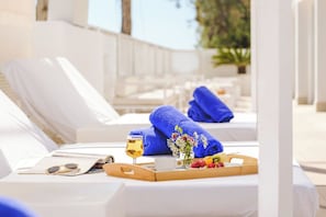 A sunlit poolside lounge area with white lounge chairs, blue towels, a tray with fruits, a glass of wine, and sunglasses.