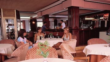 A restaurant with round tables and wicker chairs, a bar area, and a view of the kitchen.