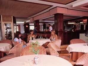 Un restaurante con mesas redondas y sillas de mimbre, una zona de bar y vistas a la cocina.