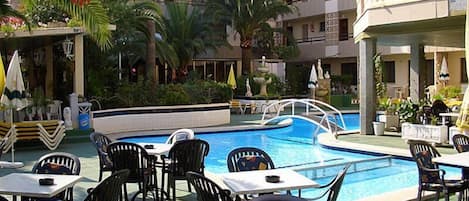 A poolside area with multiple tables and chairs, a swimming pool, and surrounding greenery.