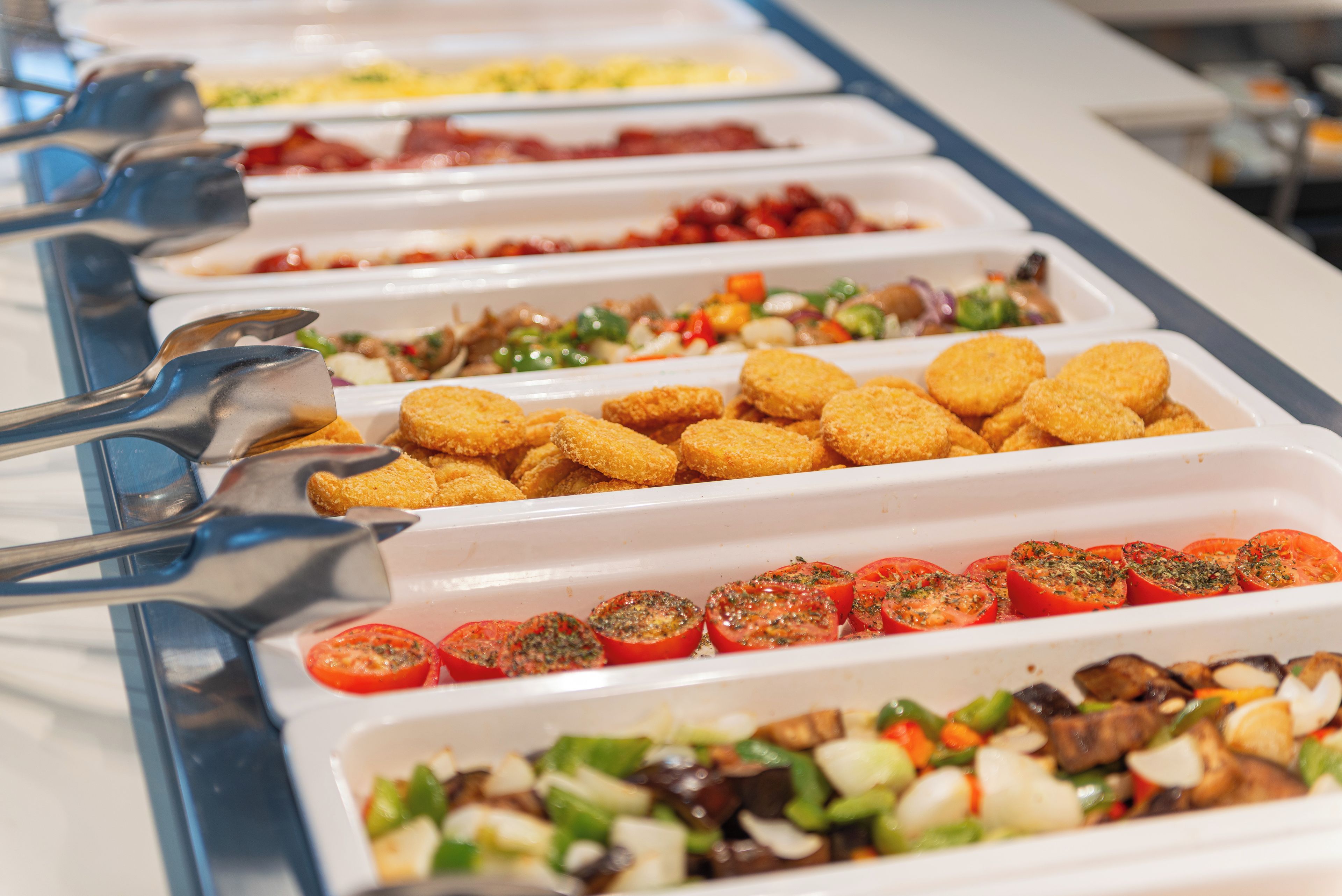 A buffet line with various food options including breaded items and sliced tomatoes.