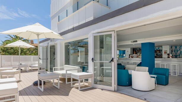 An outdoor deck with white furniture and umbrellas, a bar area with blue accents, and a glass door entrance. - Welikehotel Triton Beach (Capdepera)