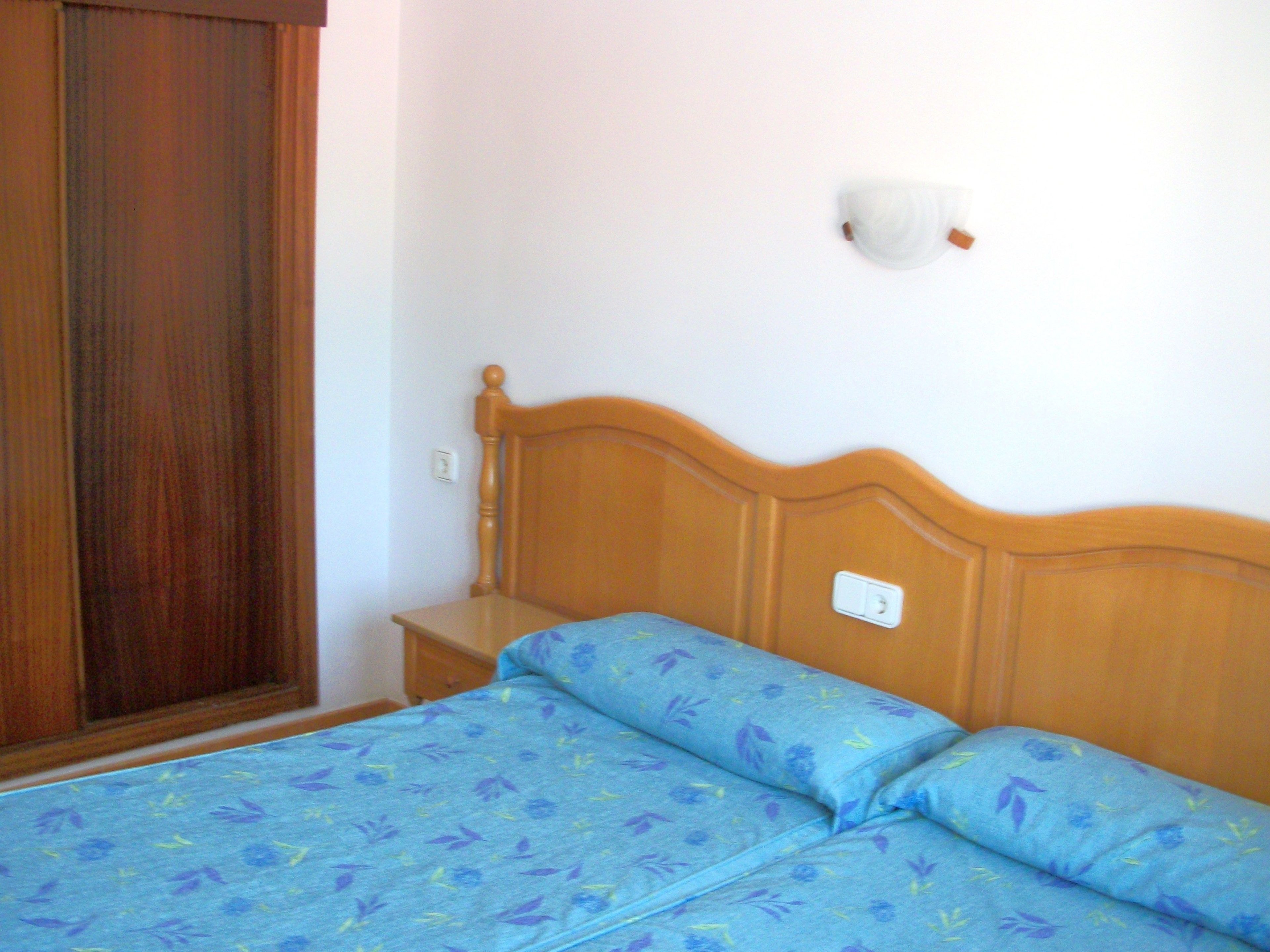 A bedroom with a wooden headboard, a bed with blue floral bedding, a nightstand, and a wall-mounted light fixture.