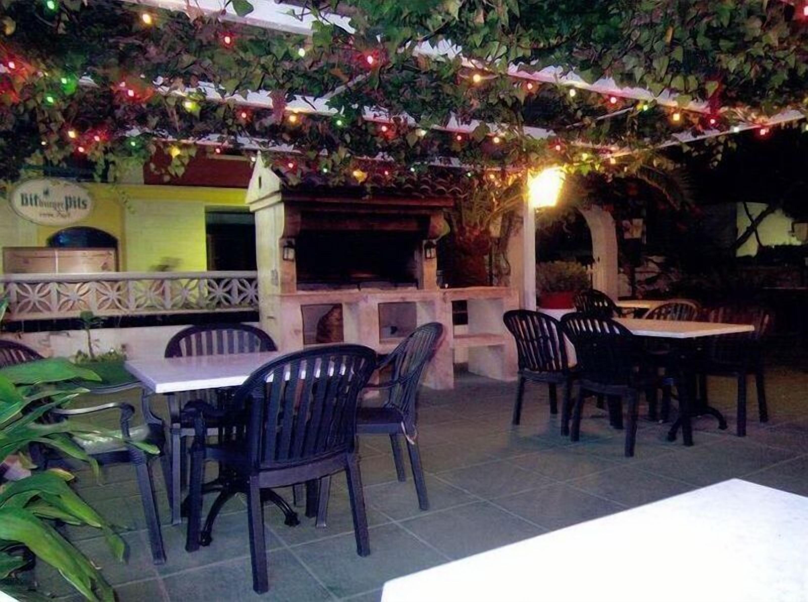 Un área de comedor al aire libre con mesas y sillas, luces colgantes y una estación de parrillada.