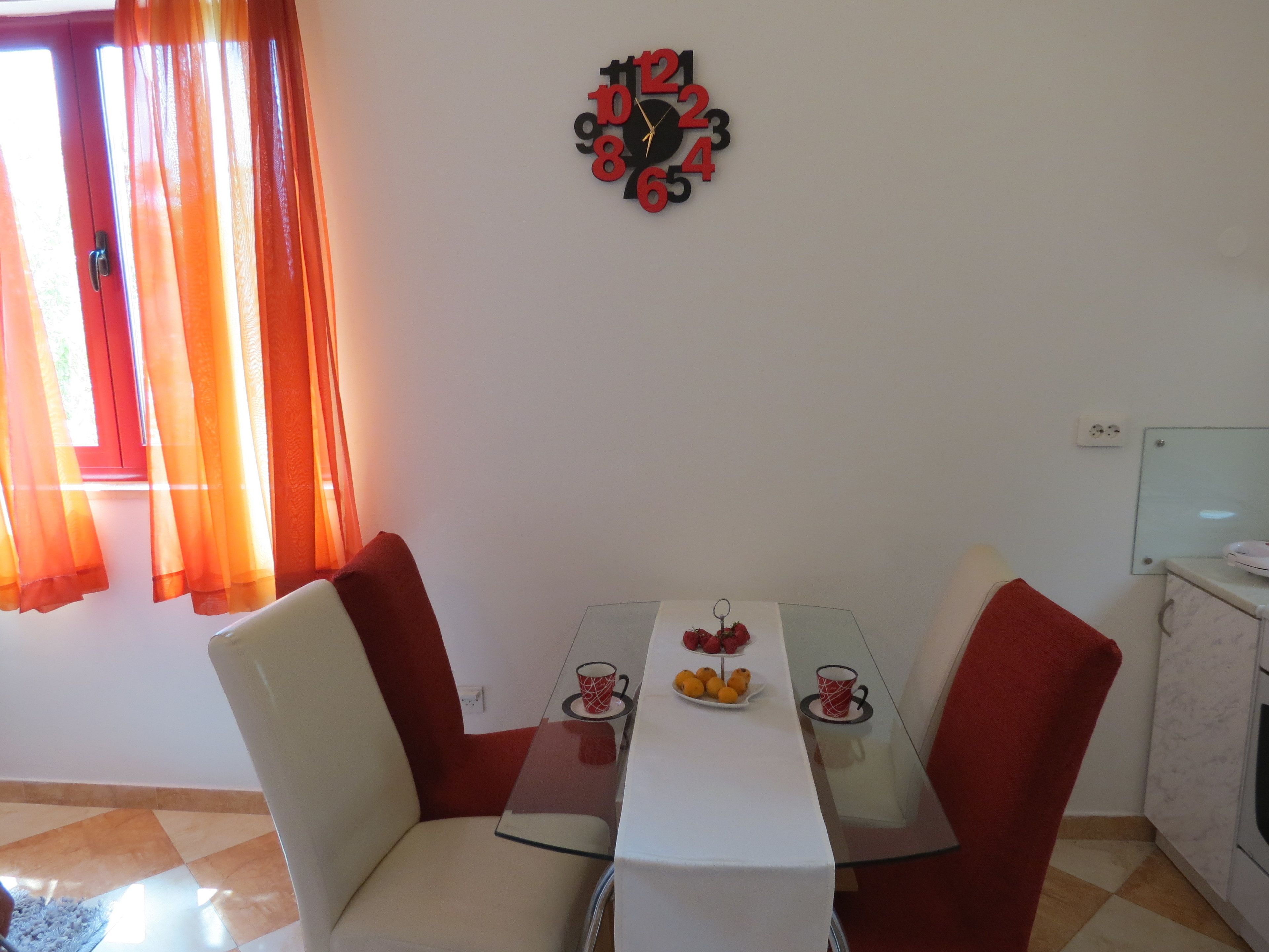 Un comedor con mesa de vidrio, dos sillas, un reloj de pared y una ventana con cortinas.
