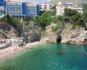 A coastal area with a beach, a rocky cliff, and buildings in the background. - Apartment Gordana (Dubrovnik)