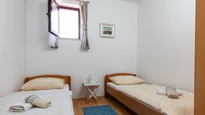 Une petite chambre propre, avec deux lits, une fenêtre avec des rideaux, une table de chevet avec une lampe et un tableau encadré au mur.