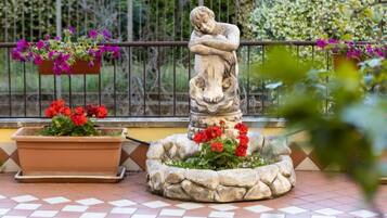 Una scultura in pietra raffigura una figura che abbraccia un bambino, circondata da un cordone di pietra e vasi con fiori rossi.