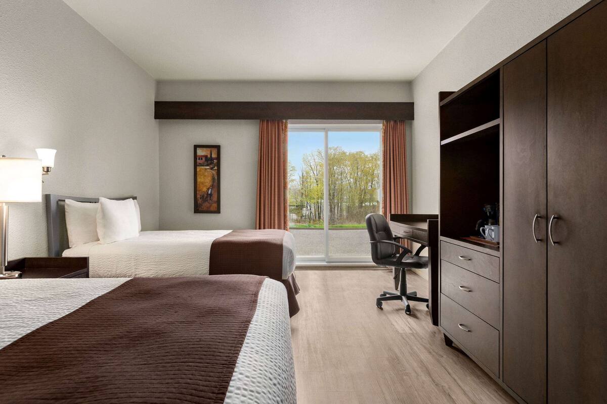 Habitación de hotel con dos camas, un escritorio, una silla, un armario y un ventanal grande con cortinas.