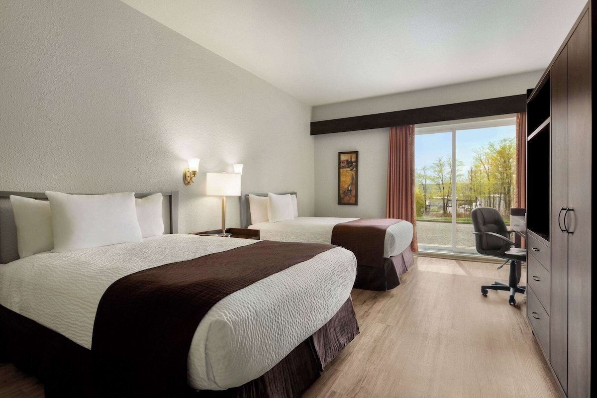 Habitación de hotel con dos camas, un escritorio, una silla y un ventanal con vistas a árboles.