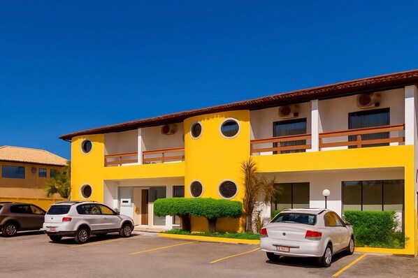 Um edifício amarelo com varanda, parque de estacionamento e dois carros estacionados em frente.
