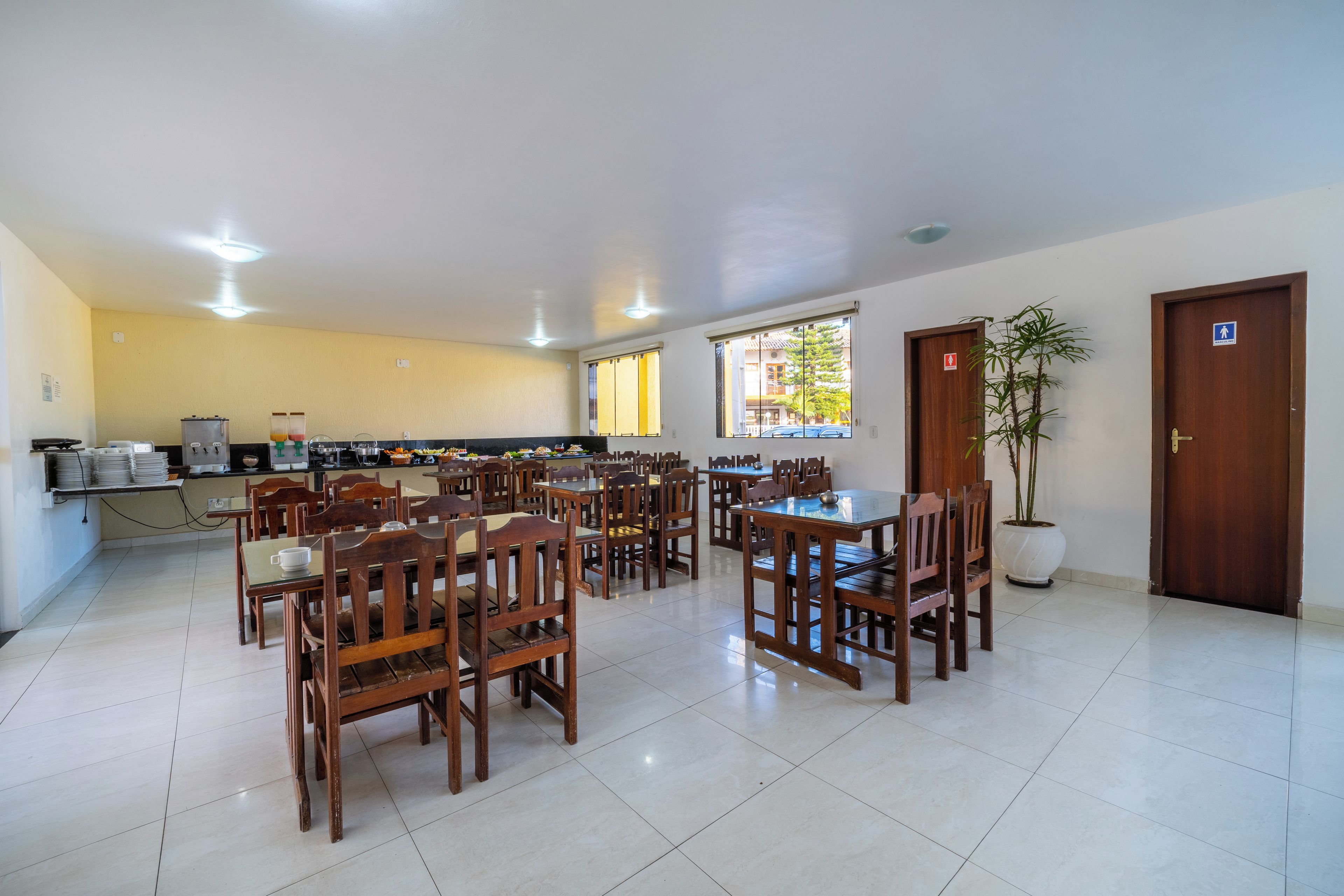 Un'ampia sala da pranzo con diversi tavoli e sedie in legno, una cucina sullo sfondo e un'ampia finestra che lascia entrare la luce naturale.