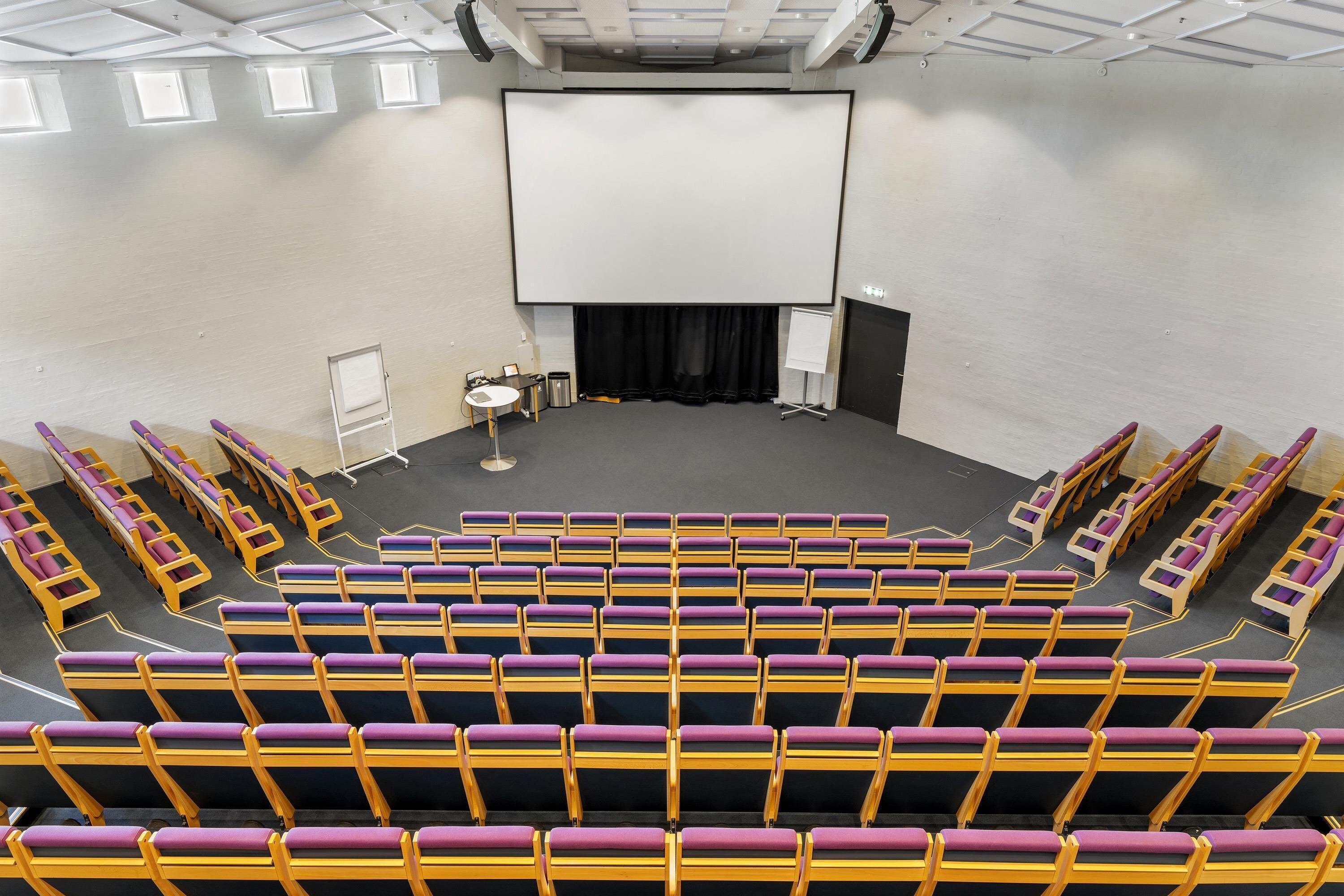 A theater with rows of yellow and purple seats, a large screen, and a stage with curtains.