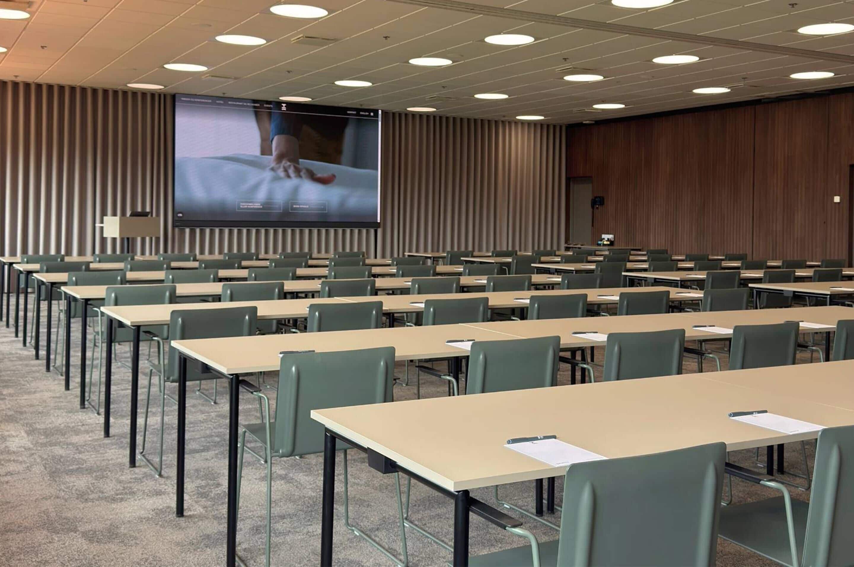 Ein Konferenzraum mit Tischen, Stühlen in Reihen, einer großen Leinwand und einer Holzwand.