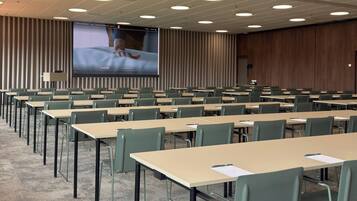 Ein Konferenzraum mit Tischen, Stühlen in Reihen, einer großen Leinwand und einer Holzwand.
