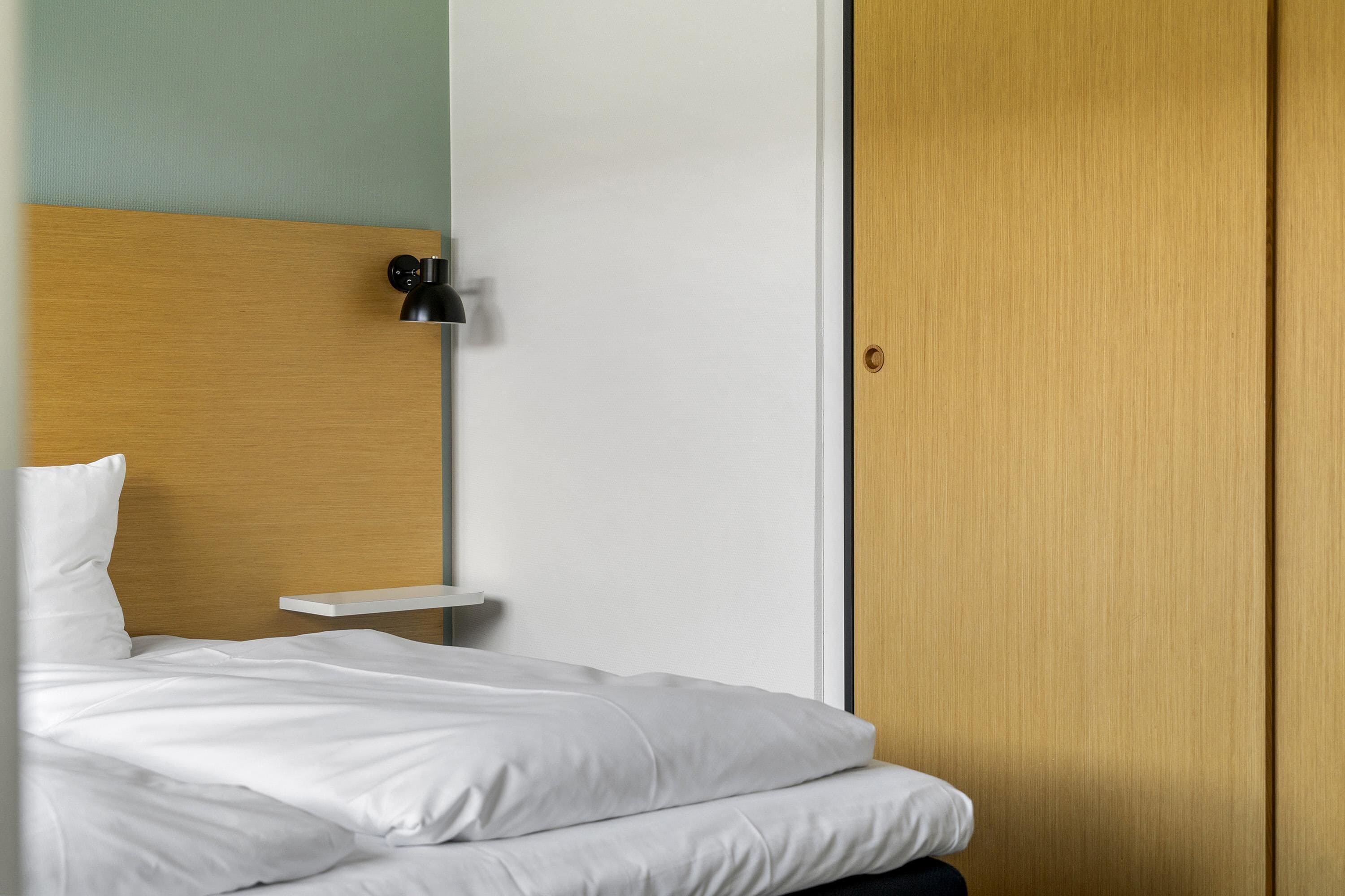 A neatly made bed with white linens, a bedside table with a lamp, and a wooden wardrobe.