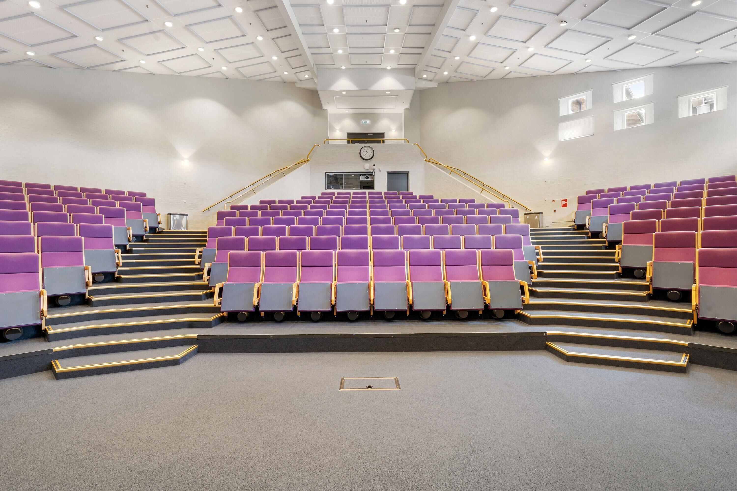 Ein großer Saal mit abgestuften Sitzreihen, einer Uhr und einer Treppe zu einer oberen Ebene.