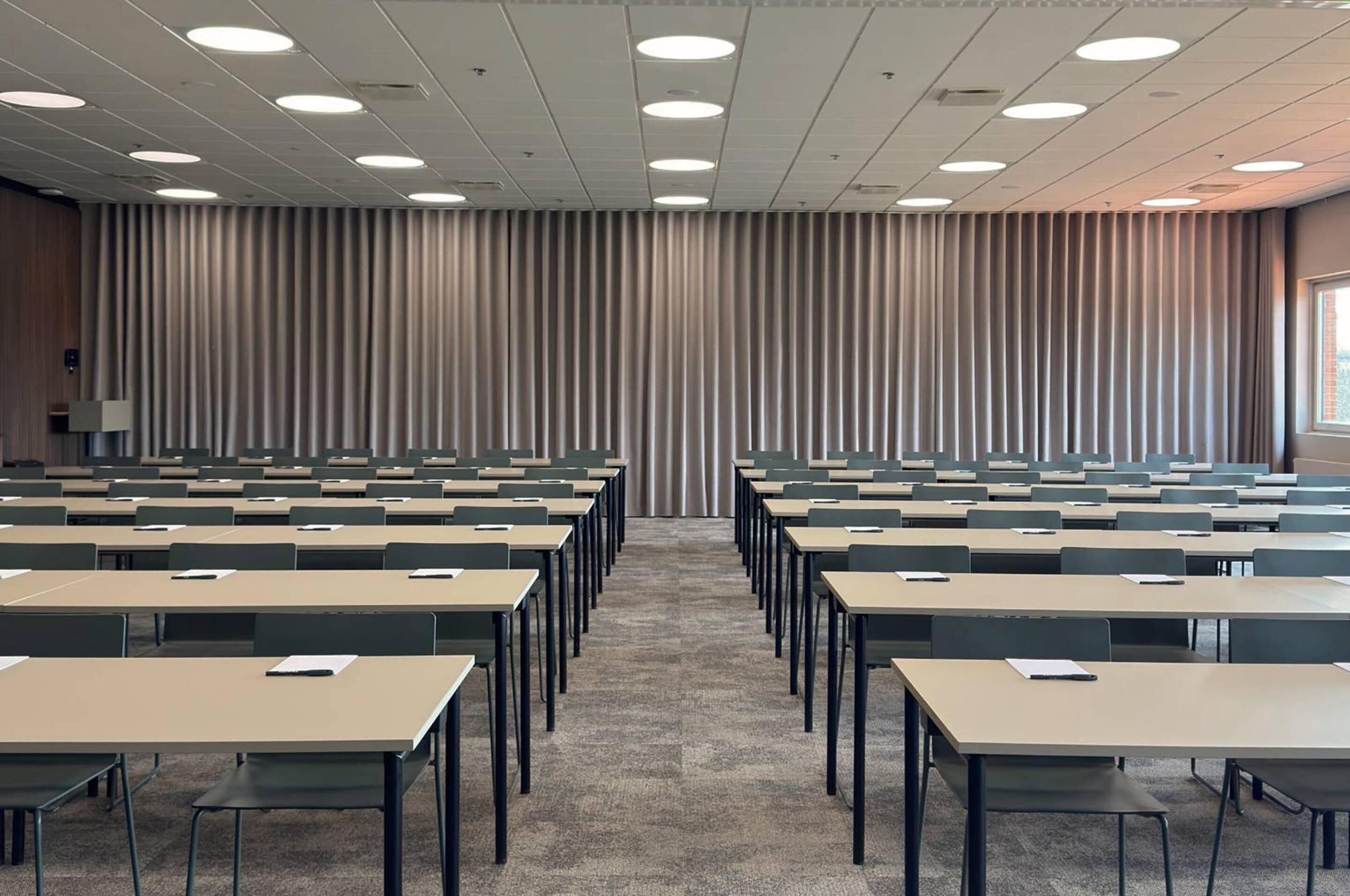 A conference room with rows of tables and chairs, a carpeted floor, and a curtain wall with vertical blinds.