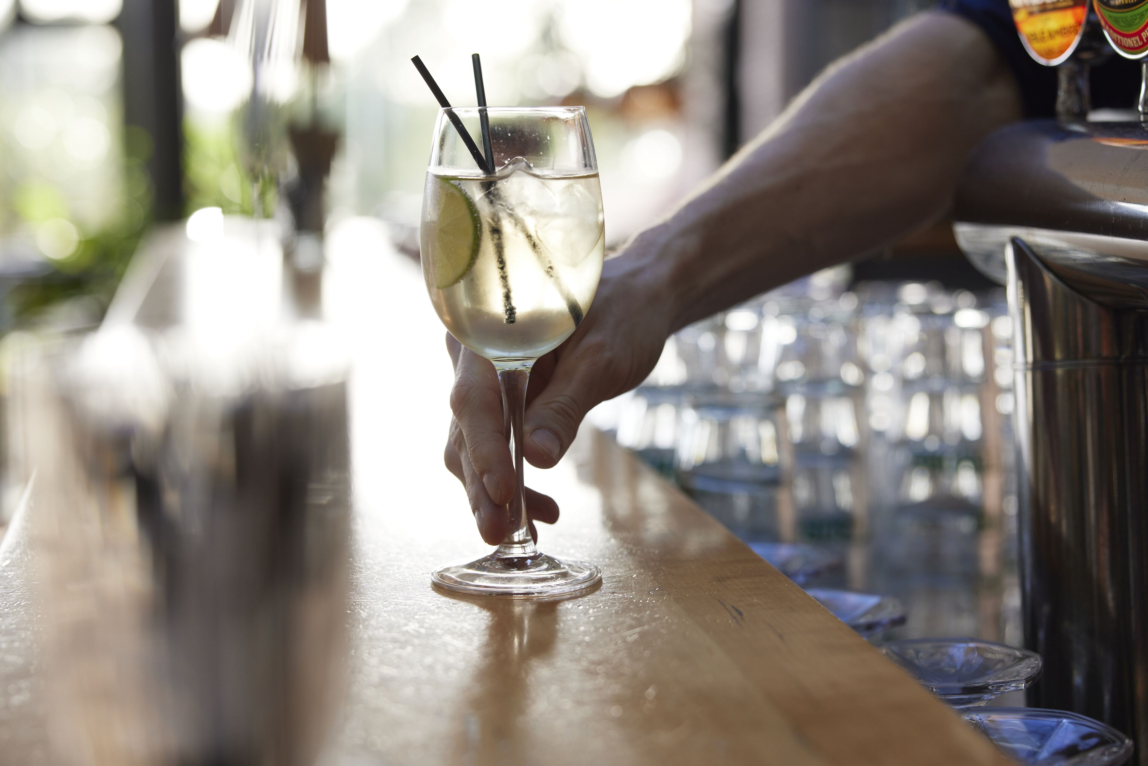 A hand holding a glass with a straw and lime slice, placed on a bar counter with glasses and a bottle in the background.