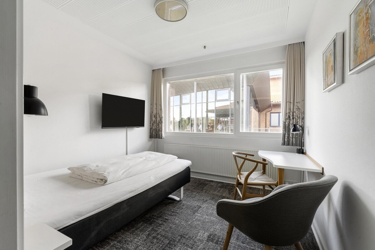 Una habitación de hotel moderna con cama, escritorio, silla, televisor y un ventanal grande.