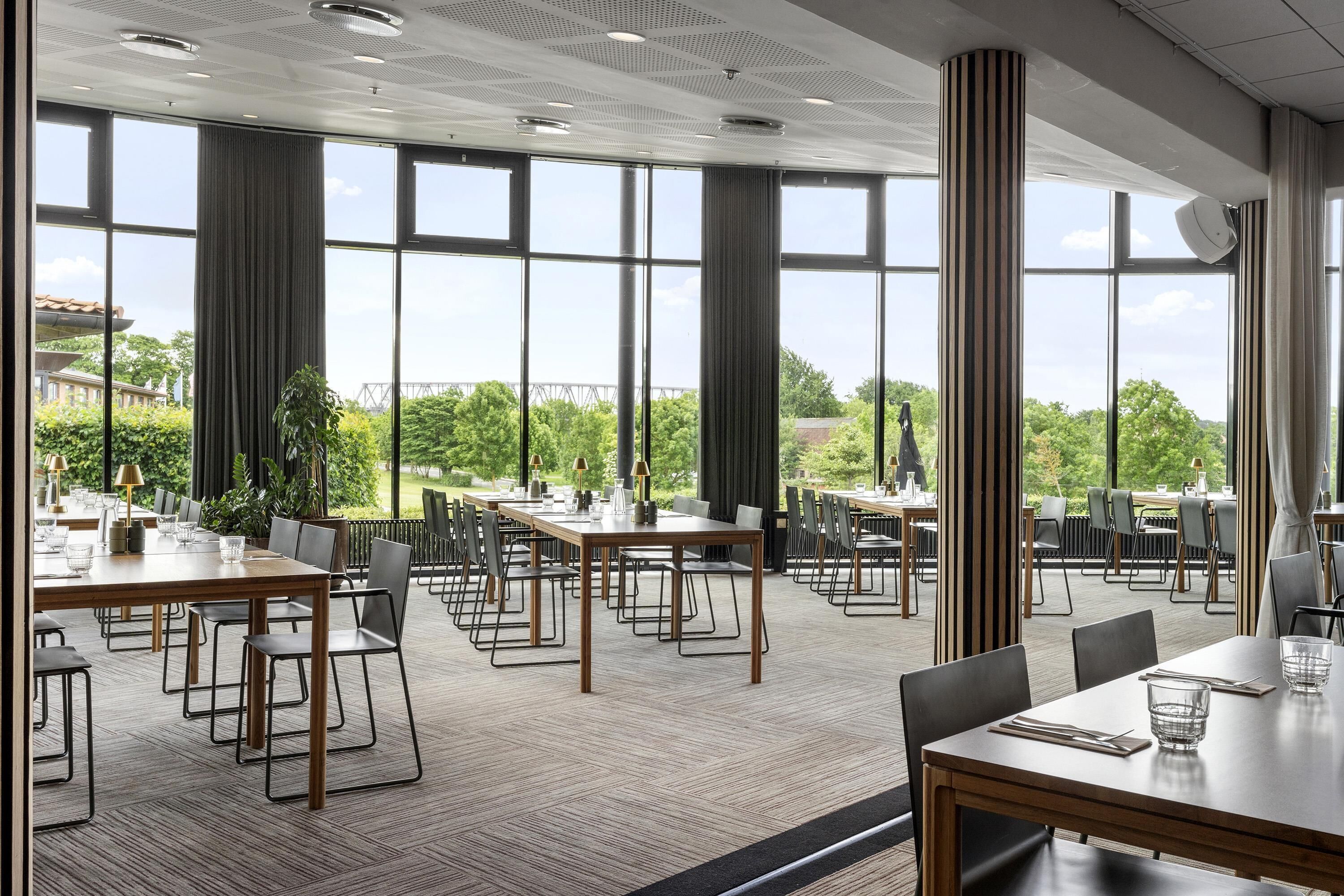 A spacious dining area with multiple tables and chairs, large windows, and a view of greenery outside.