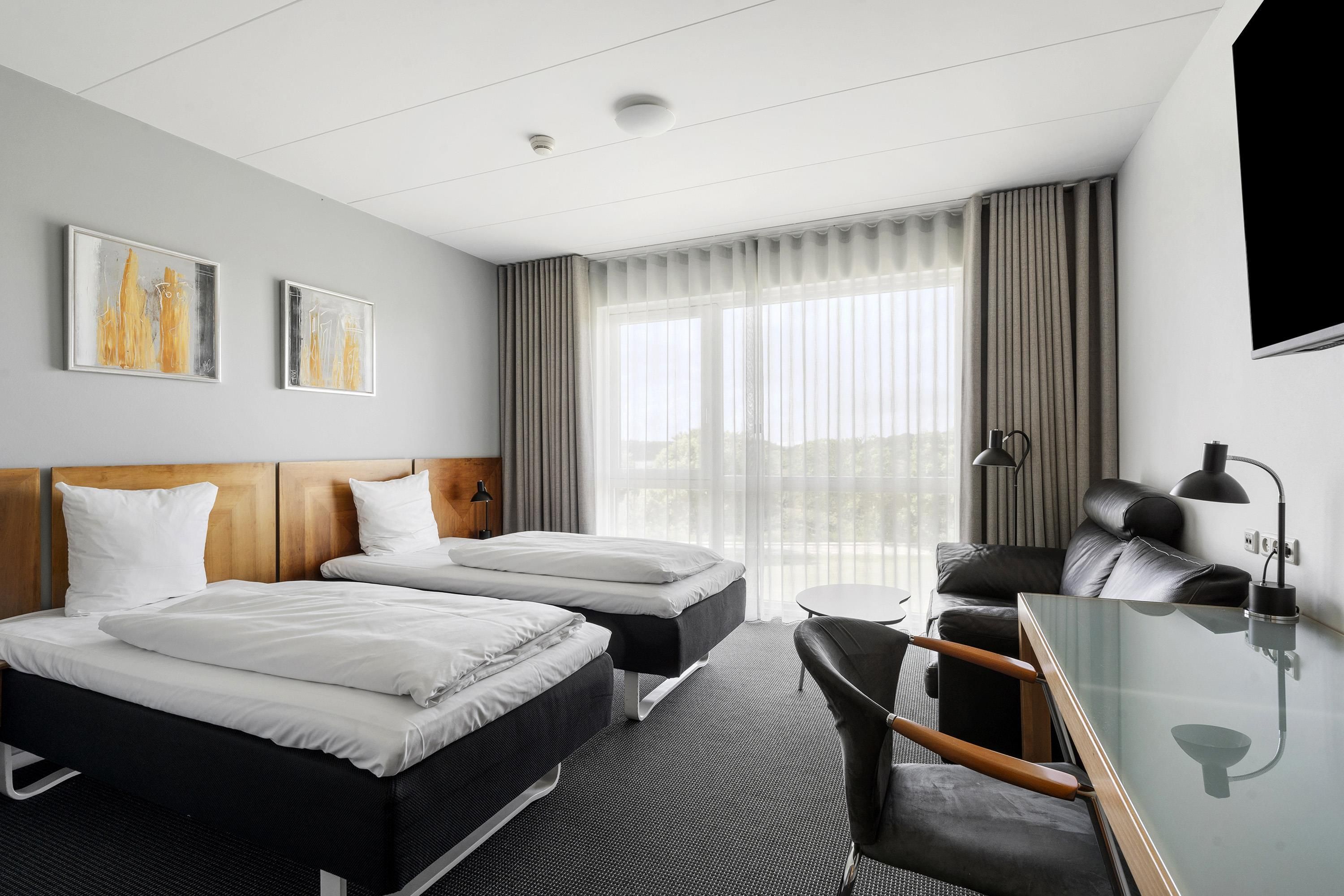 A hotel room with two beds, a desk, a chair, a large window with curtains, and two wall art pieces.