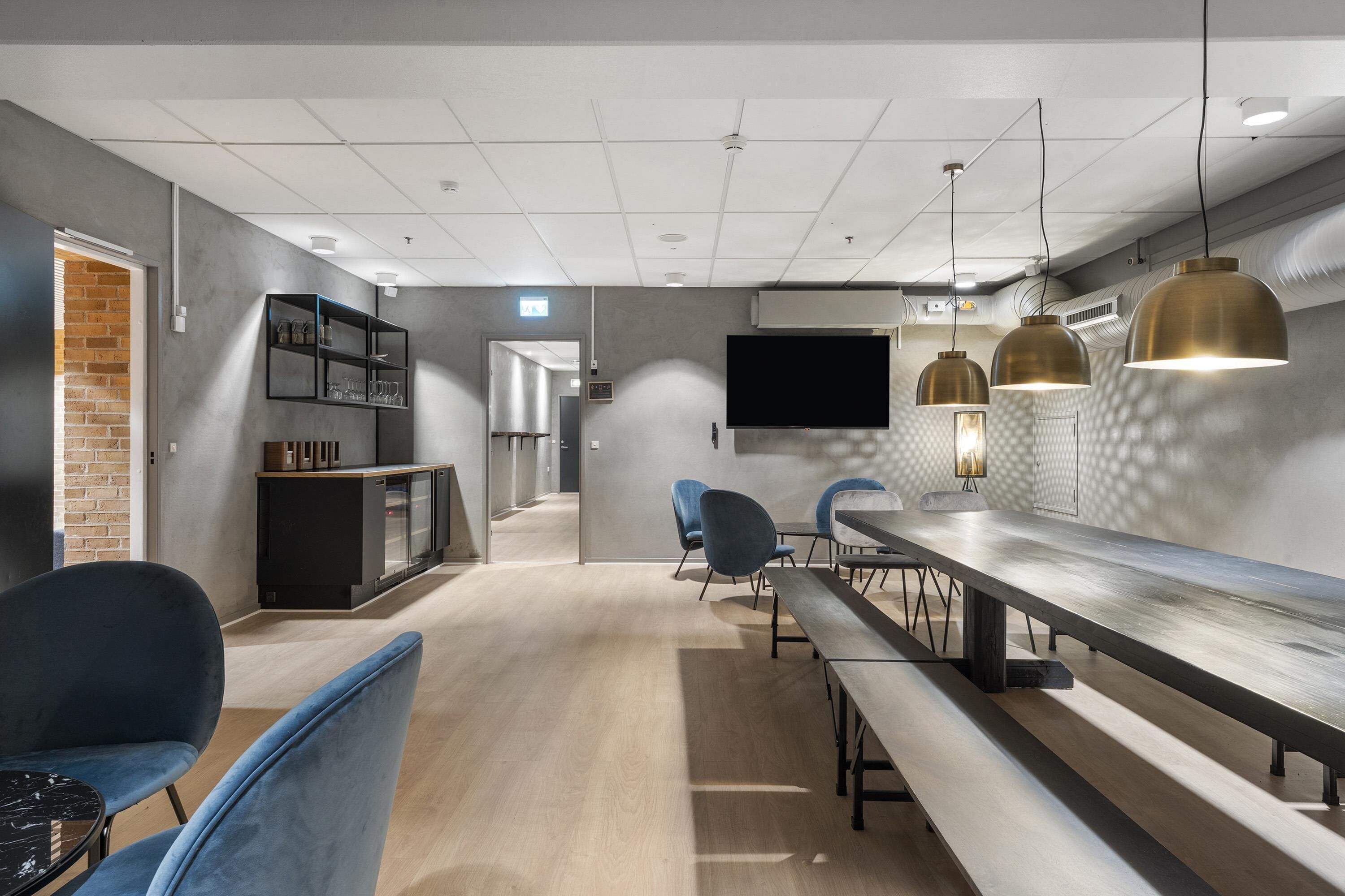 A modern interior with a long dining table, blue chairs, a TV, pendant lights, and a brick wall.