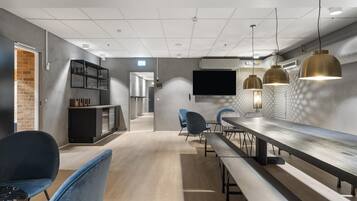 A modern interior with a long dining table, blue chairs, a TV, pendant lights, and a brick wall.